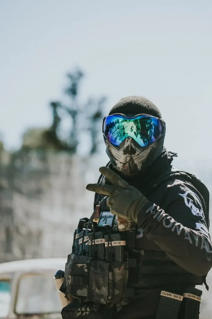 A paintball player in full gear making a peace sign during an outdoor game.